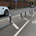 Milestone islands installed along a city street, featuring a continuous line of slim modular bases with reflective bollards that separate traffic lanes, guiding vehicles past pedestrians and urban buildings.
