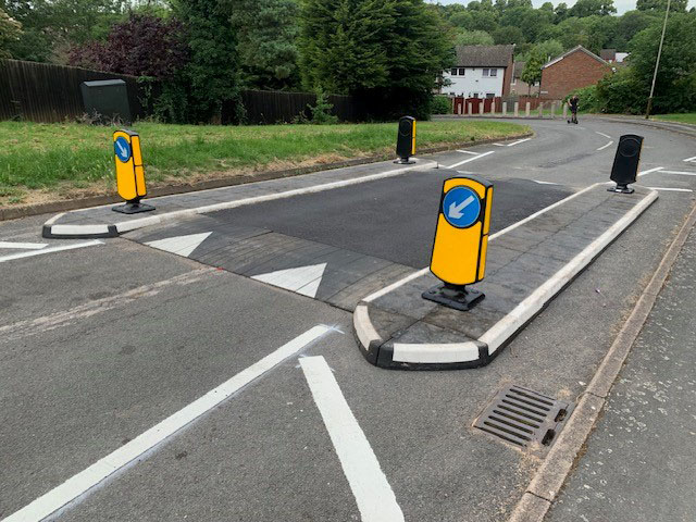Angled view of a raised speed table combined with RediPave traffic islands, showing black and white modular island units with yellow keep-left bollards separating traffic lanes on a residential street.