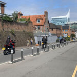 Milestone islands installed along a city street, featuring a continuous line of slim modular bases with reflective bollards that separate traffic lanes, guiding vehicles and motorcycles past pedestrians and urban buildings.