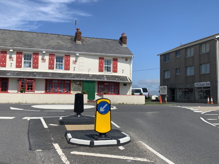 RediPave pedestrian refuge island installed at a road junction, featuring a modular traffic island base with integrated black and white markings and keep-left bollards to guide vehicles and provide pedestrian protection.