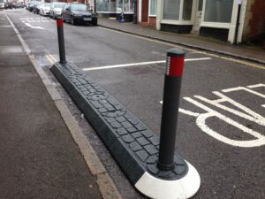 Satellite traffic island installed in the carriageway, formed with modular kerbing and fitted with black bollards to guide vehicles and control traffic flow.