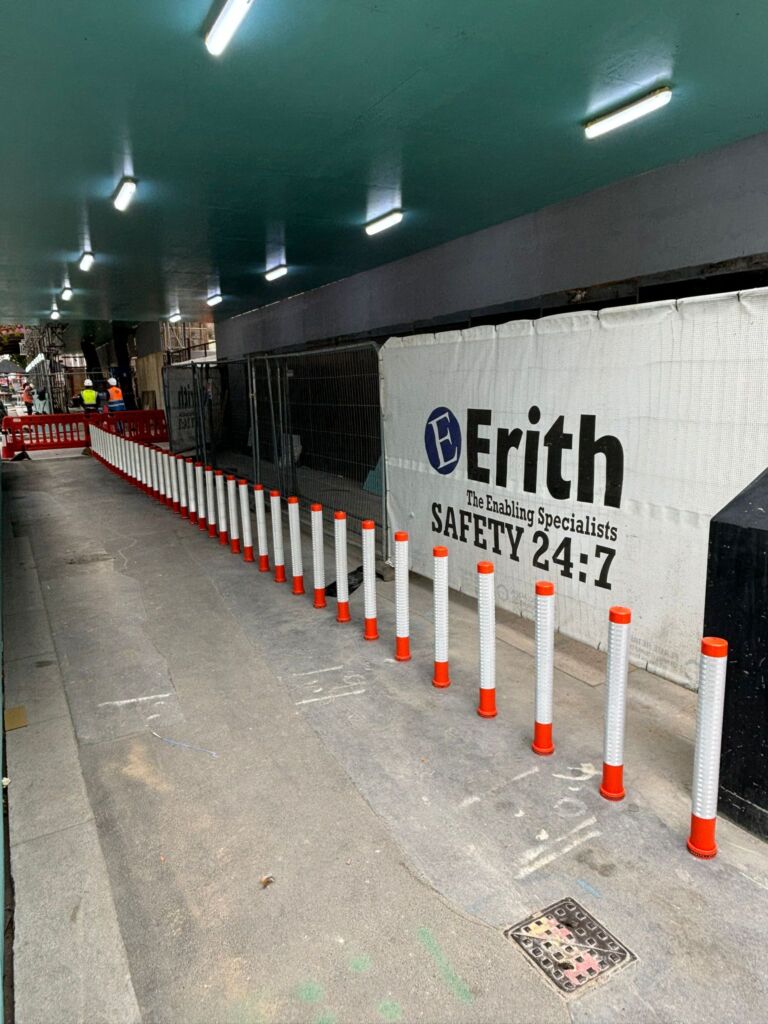 On-highway orange and white pole cones forming a safety barrier to protect pedestrians and cyclists along a covered footway beside construction works