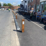 Milestone island installed along the edge of a footway, featuring modular kerb units with integrated light domes and timber verge posts, separating the carriageway from outdoor seating along a street.