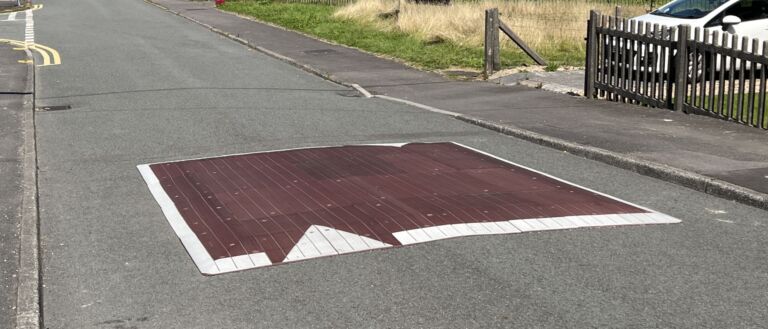 Raised rubber speed table installed on a residential road, with contrasting surface and road markings to slow vehicle traffic near a pedestrian area.