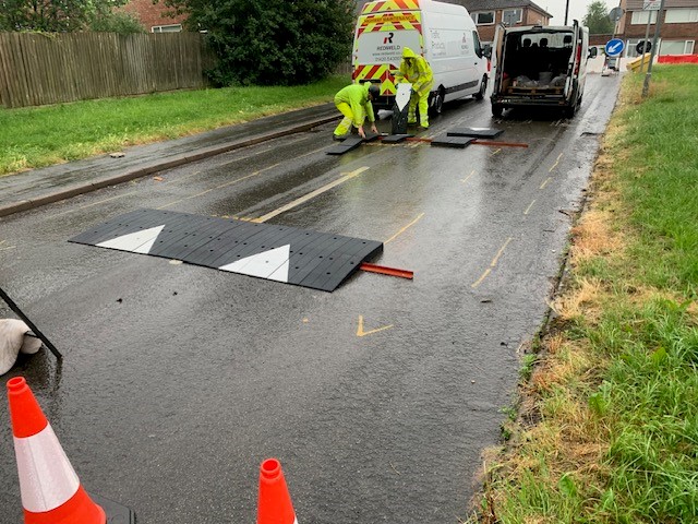 Road crew installing modular speed table on residential street in wet conditions, with traffic cones and vans nearby