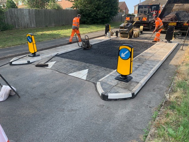 Workers compacting newly laid asphalt within a traffic island speed table, with bollards and kerbed islands in place