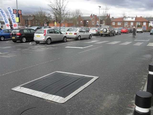 Black and white speed cushion used for traffic calming in busy retail car park with parked vehicles.