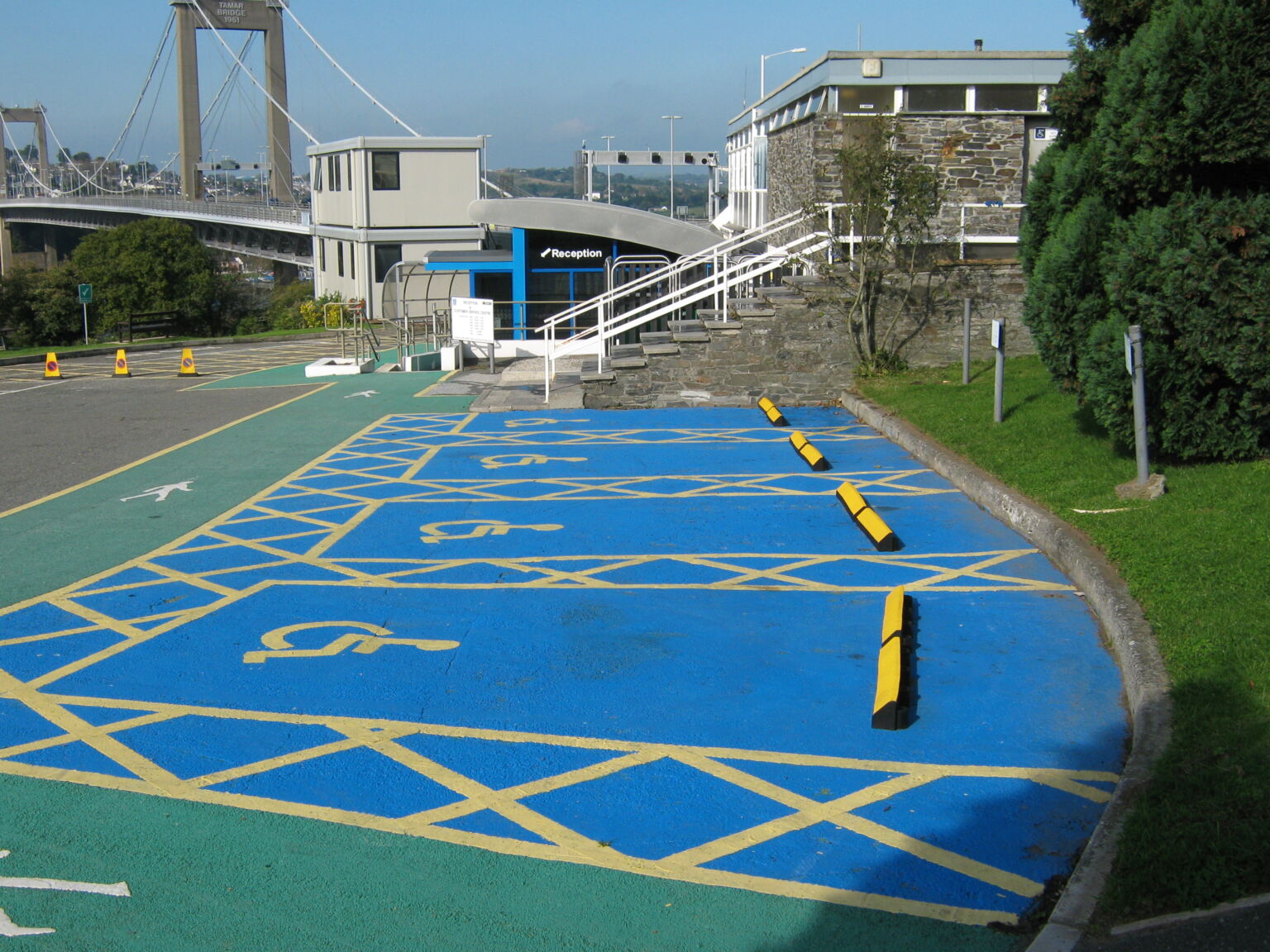 Accessible parking bays with blue markings and wheelchair symbols outside building reception, with bridge visible in background.