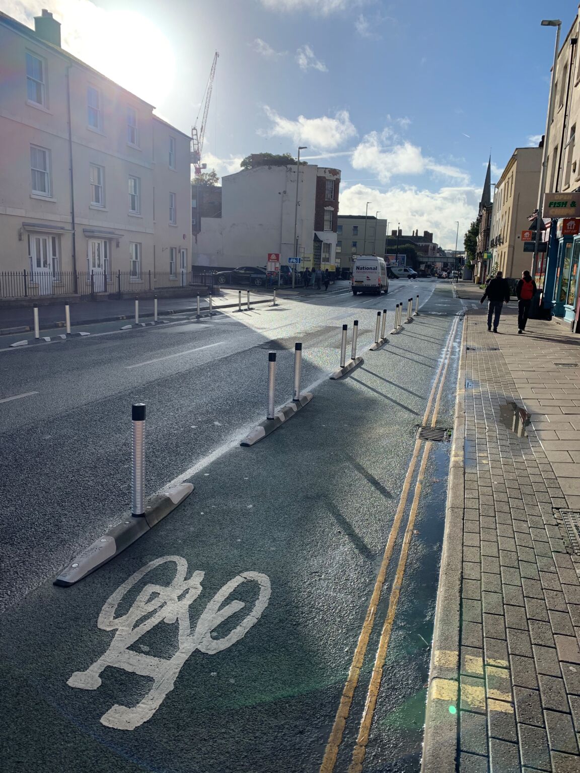 Empty segregated cycle lane with continuous row of flexible bollards alongside shops on urban road.