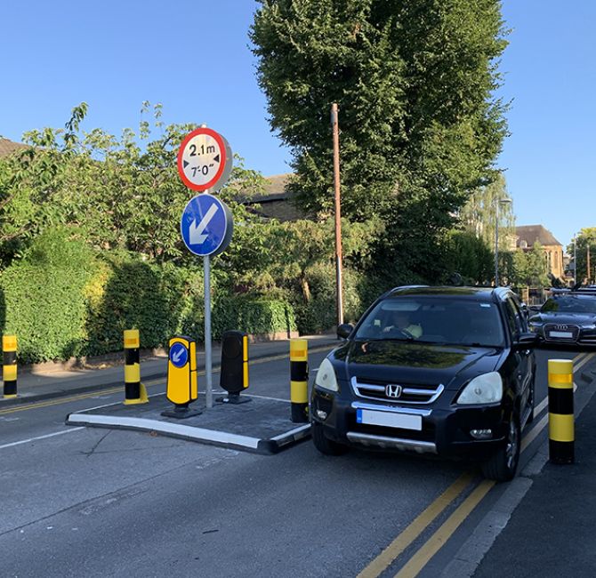 RediPave traffic island installed on an urban street, featuring a modular island base with integrated black and white markings, reflective bollards, and a keep-left sign, positioned alongside parked vehicles to manage traffic flow.