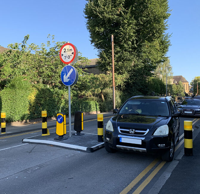 RediPave traffic island installed on an urban street, featuring a modular island base with integrated black and white markings, reflective bollards, and a keep-left sign, positioned alongside parked vehicles to manage traffic flow.